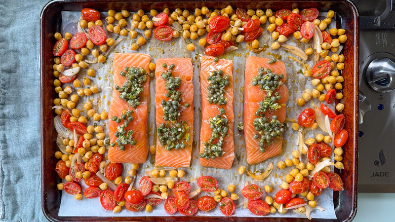 Raw salmon filets on baking sheet with caper topping and chickpeas, grape tomatoes, and shallot