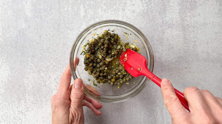 Mixing capers, lemon, garlic, and rosemary with olive oil in glass bowl with rubber spatula