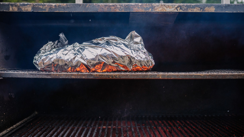 Meat wrapped in tin foil smoking on top rack of a grill