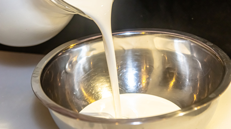 A glass jar of heavy cream pouring into a stainless steel bowl