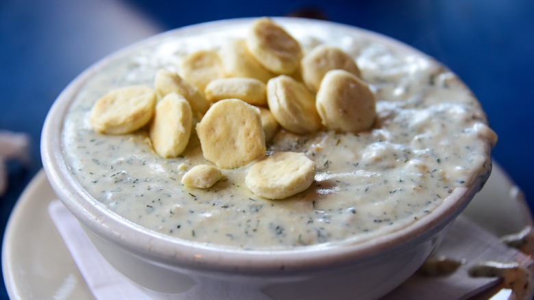 A white bowl of clam chowder on a white saucer with oyster crackers on top in front of a blue background