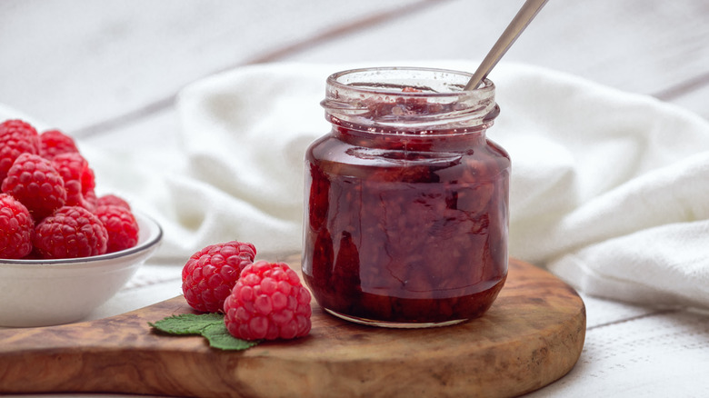 A jar of raspberry jam and fresh raspberries are arranged on a table with a white background.
