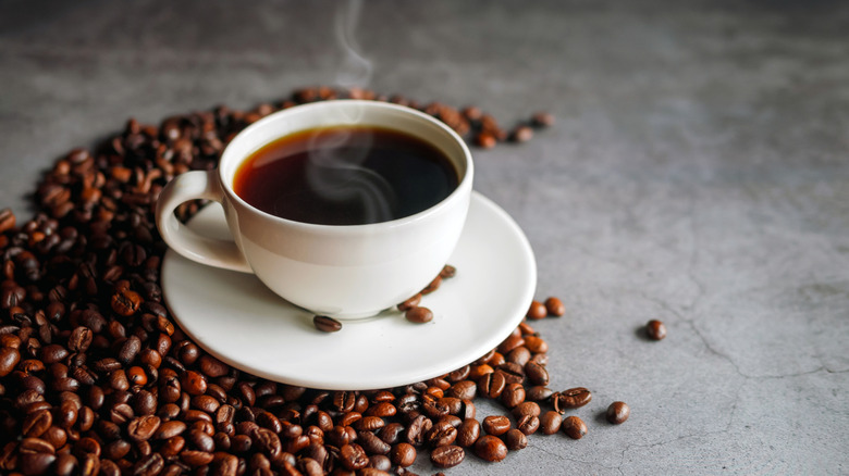 A steaming cup of coffee is placed on coffee beans on a gray table.