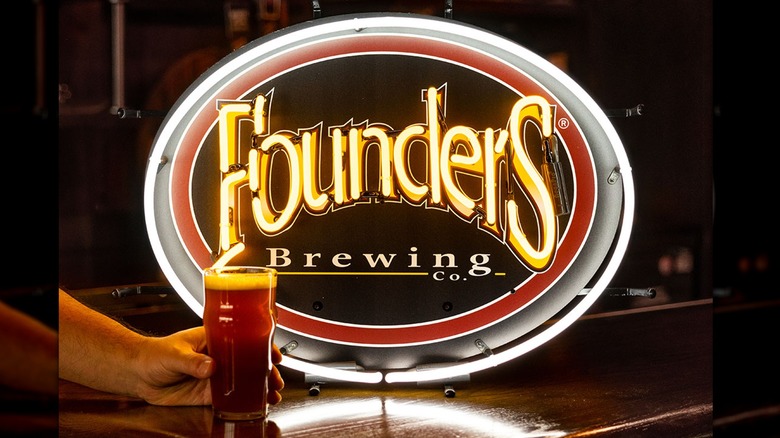 A person holding a glass of ale in front of the Founders Brewing Company sign