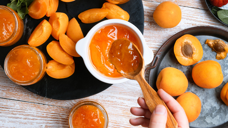 woman spooning apricot jam into container on table full of fresh apricots