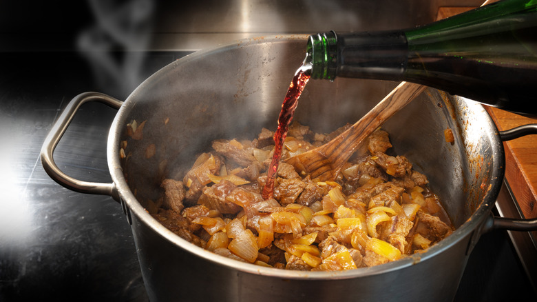 red wine being poured into a pot of cooking beef to deglaze