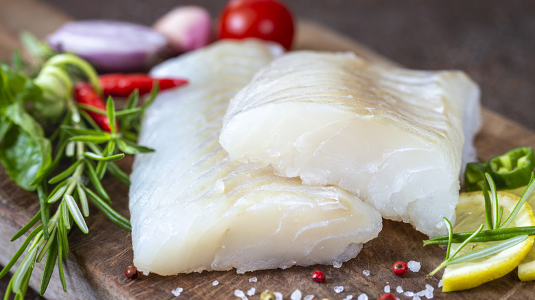 Cod fillet on wooden chopping board with vegetables, herbs, and seasonings.