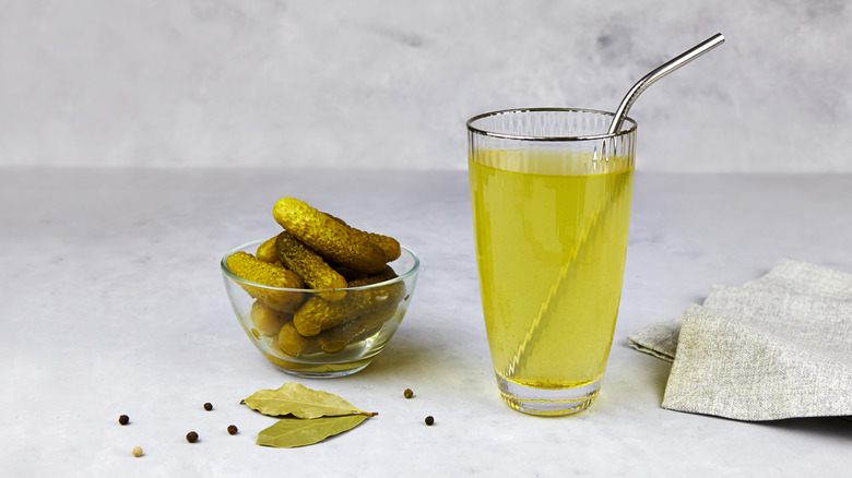 A glass of pickle juice with a metal straw next to a small glass bowl of mini pickles