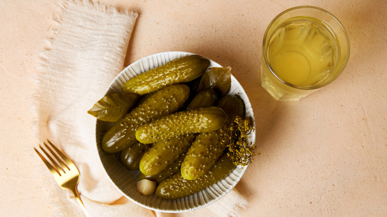 A white bowl of pickles and a glass of pickle juice with a gold fork and cloth napkin on a beige background