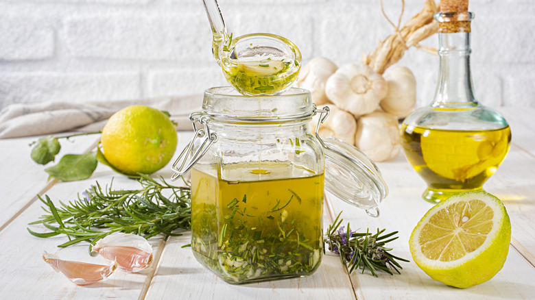 A ladle taking dressing from a jar of oil, vinegar, herbs, garlic, and lemon surrounded by ingredients