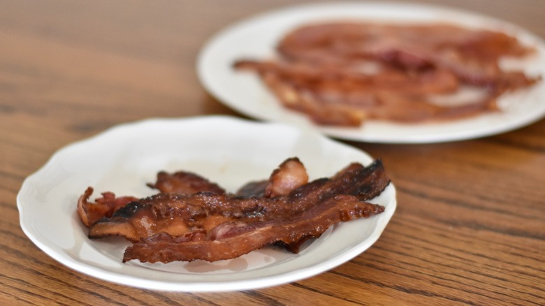 Six slices of crispy bacon on a white plate with another larger plate of bacon in the background on a wooden table