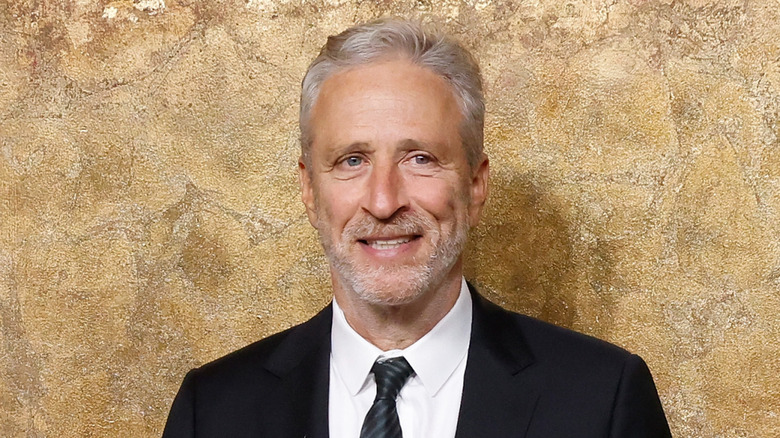 John Stewart standing in front of a gold wall and wearing a dark suit and tie