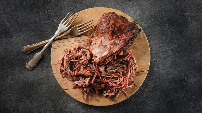 A wooden circular cutting board with pulled pork on it and two forks against a dark gray background