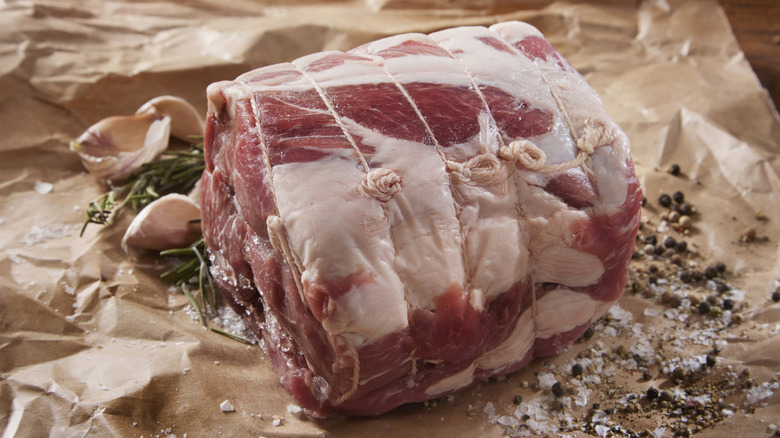 A raw pork shoulder with fat on the outside wrapped in string on a parchment paper with salt, pepper, and garlic