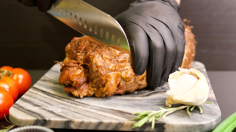 A person cuts into a smoked pork shoulder with a knife on a marble cutting board wearing black gloves with garlic, tomatoes, and herbs nearby