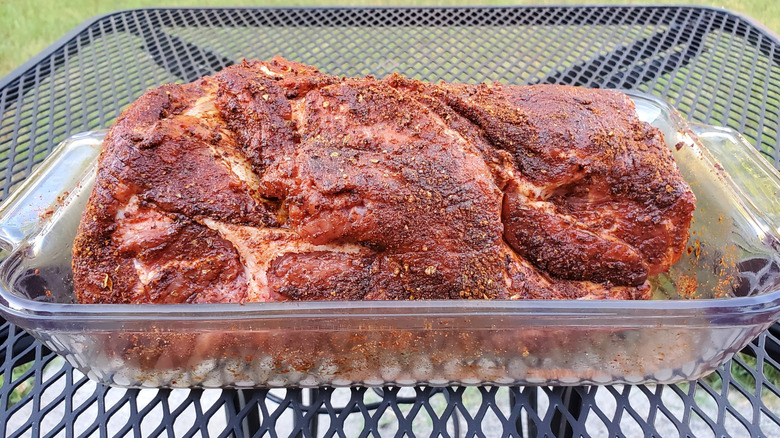 A pork shoulder in a glass dish with seasoning rub on the outside on a metal grate table