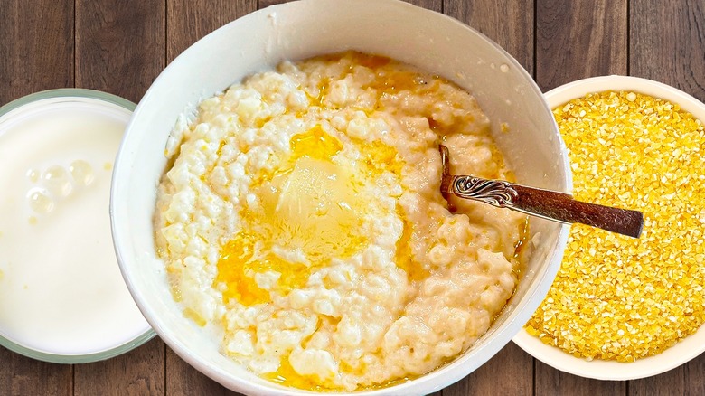 A bowl of grits with a spoon in it is flanked by containers of raw grits and cream.