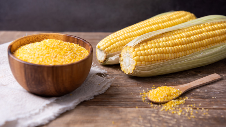 A bowl of coarse uncooked grits sits next to two ears of corn.