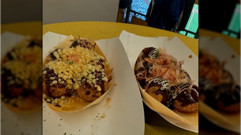 Two trays of takoyaki with different sauces and toppings like bonito flakes, on a table