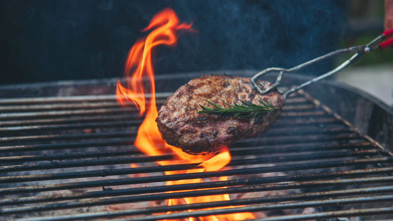 A burger being held by tongs over a grill, with flames licking the burger