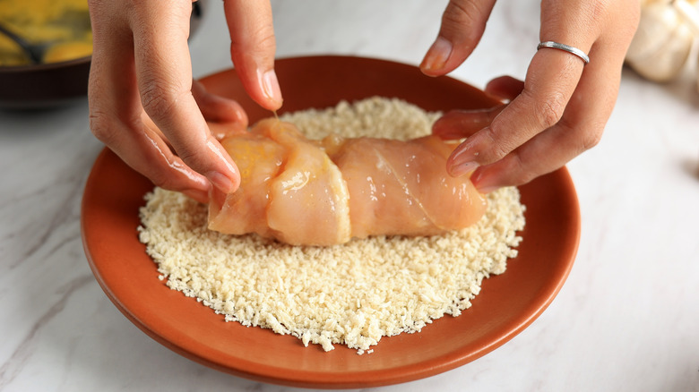 Hands coating raw chicken with bread crumbs