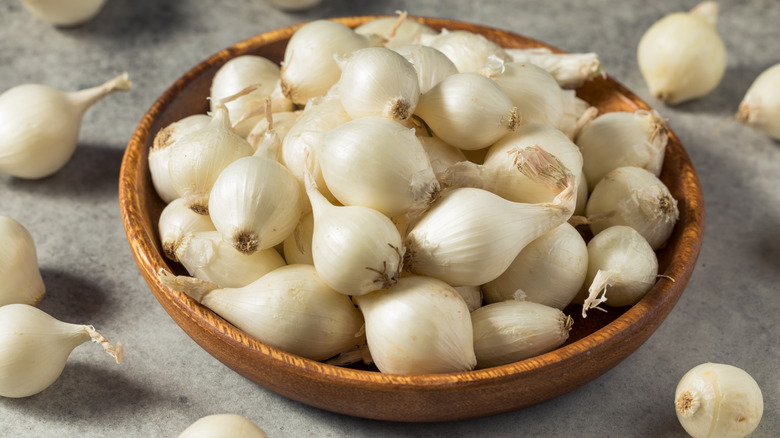 A brown bowl of raw pearl onions on a gray countertop with pearl onions scattered around