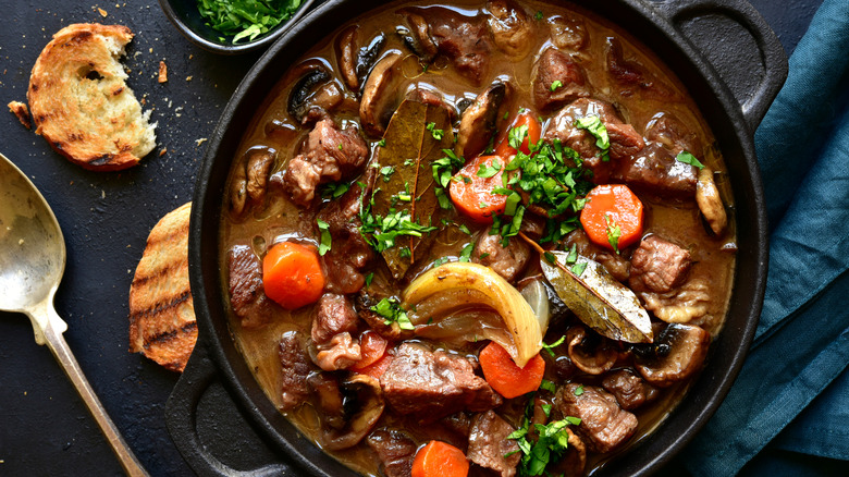 A black cast iron skillet of beef bourguignon with crusty bread and a spoon on a black counter with a blue cloth napkin