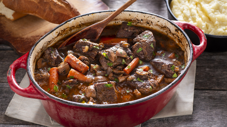 A red Dutch oven of beef bourguignon stirred by a wooden spoon on a wooden table with crusty bread and mashed potatoes in the background