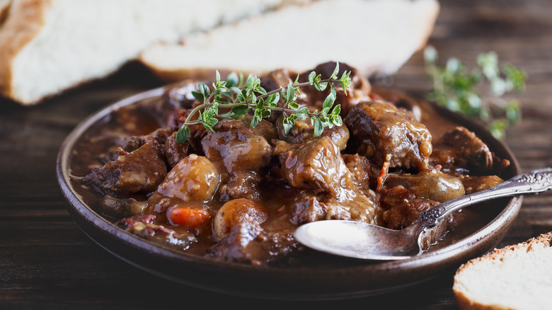 A brown bowl of beef bourguignon with a silver spoon on a wooden table with sliced bread in the background