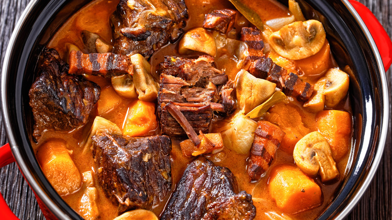 A red pot of beef bourguignon with browned meat, carrots, and mushrooms in red broth