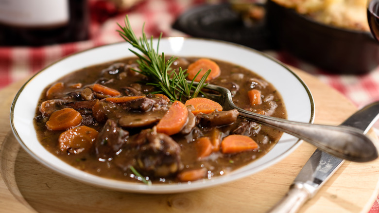 A white bowl with a gold rim of beef bourguignon with a fork and herb garnish on a wooden board on a red checkered tablecloth