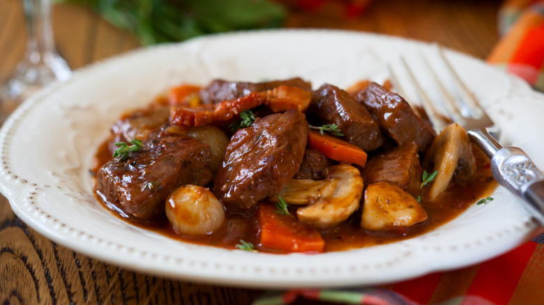 A white bowl with beef bourguignon and a silver fork on a wooden table