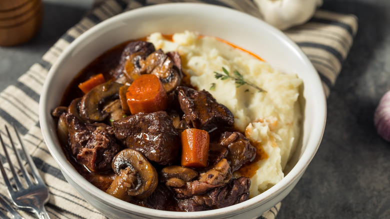 A white bowl with beef bourguignon and mashed potatoes on top of a striped cloth napkin with a fork nearby on a gray counter