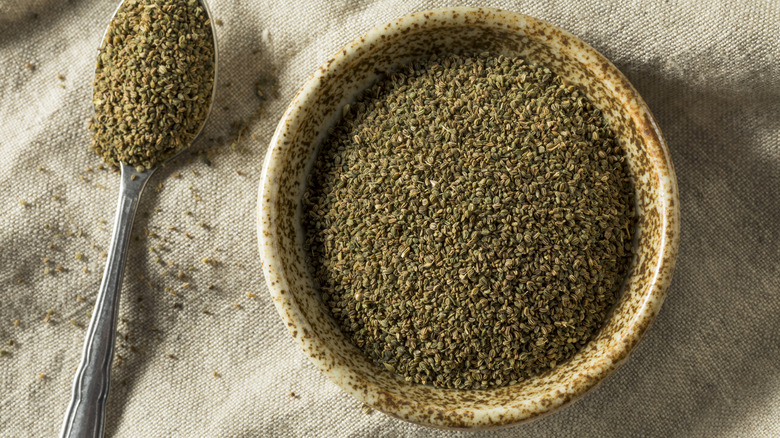 A brown bowl of ground celery seeds with a spoon full of seeds on a tan cloth background