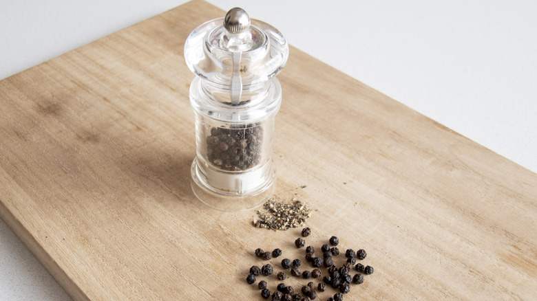 A pepper mill with whole peppercorns and ground pepper on a wooden cutting board