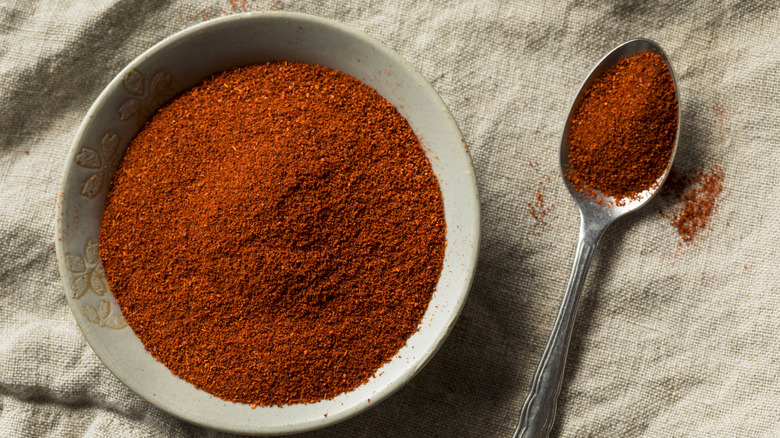 A bowl of smoked paprika powder with a spoon next to it full of a scoop of powder on a tan cloth background