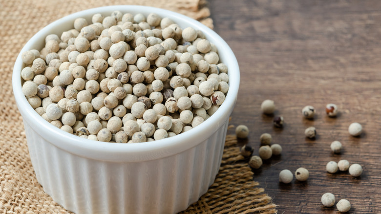 A white bowl of whole white peppercorns on a wooden counter with a burlap cloth underneath