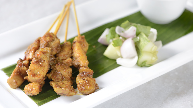 A white plate with chicken satay on a leaf