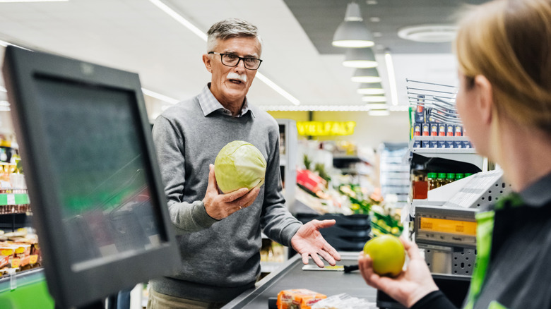 A senior male with glasses holding a cabbage as he's standing at the checkout.