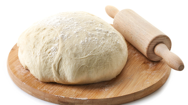 A mound of pizza dough sits on a wooden cutting board with a rolling pin