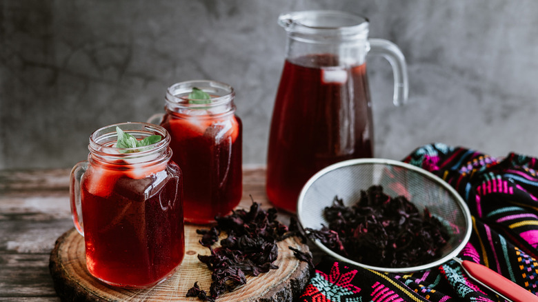 Glasses of hibiscus aguas frescas next to a jug and a strainer with hibiscus leaves