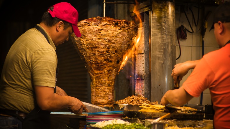 A person shaving meat off a trompo at a tacos al pastor stand