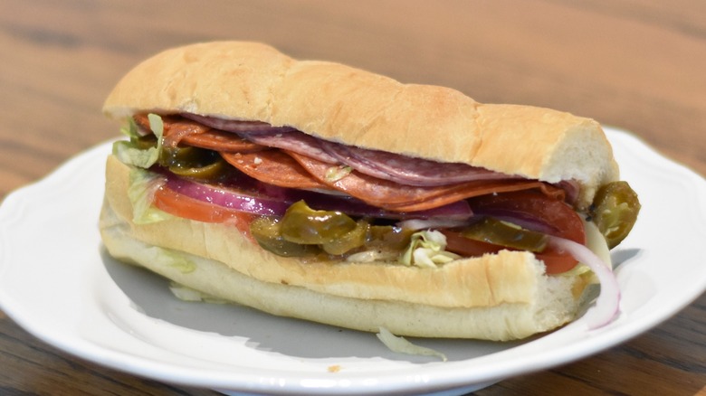 A cold cut sandwich with salami and pepperoni, topped with jalapeño, tomato, red onion, and lettuce on a white plate