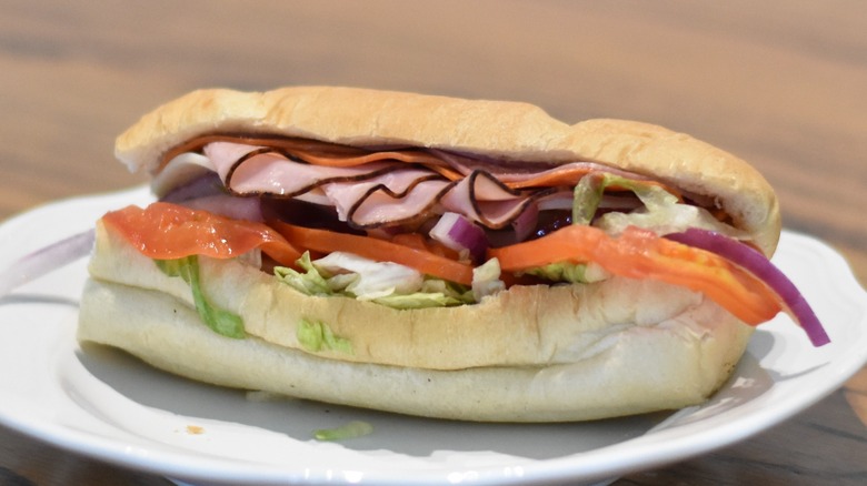 A cold cut sandwich with ham, salami, and pepperoni topped with tomato, lettuce, and red onion on a white plate