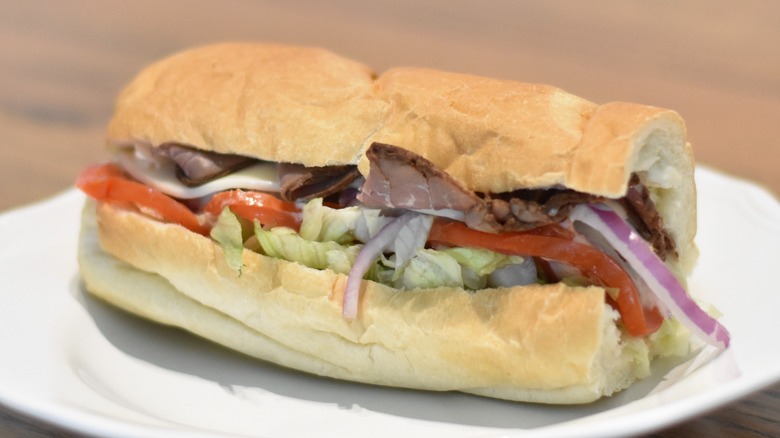 A roast beef sandwich with tomato, lettuce, cheese, and red onion on a white plate