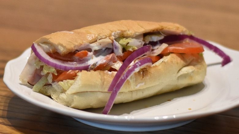 A sub sandwich with red onion, tomato, lettuce, and cold cuts on a white plate