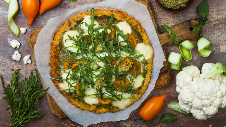 An artisinal pizza on a countertop with zucchini, squash blossoms, tomatoes, pesto, and arugula