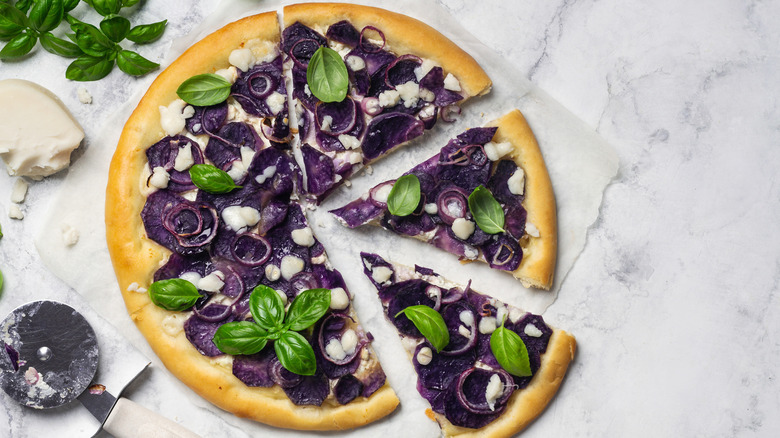 A pizza with purple potatoes, cheese, and basil on a cutting board