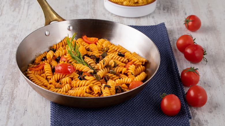 Fusilli with tomatoes and anchovies in a steel pan on a blue placemat with six cherry tomatoes with stems to its right