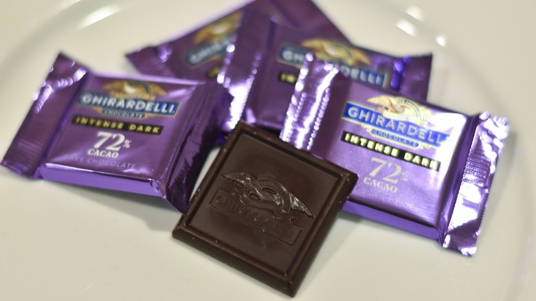 A square of Ghiradelli chocolate on a white plate next to wrapped squares of chocolate in purple foil